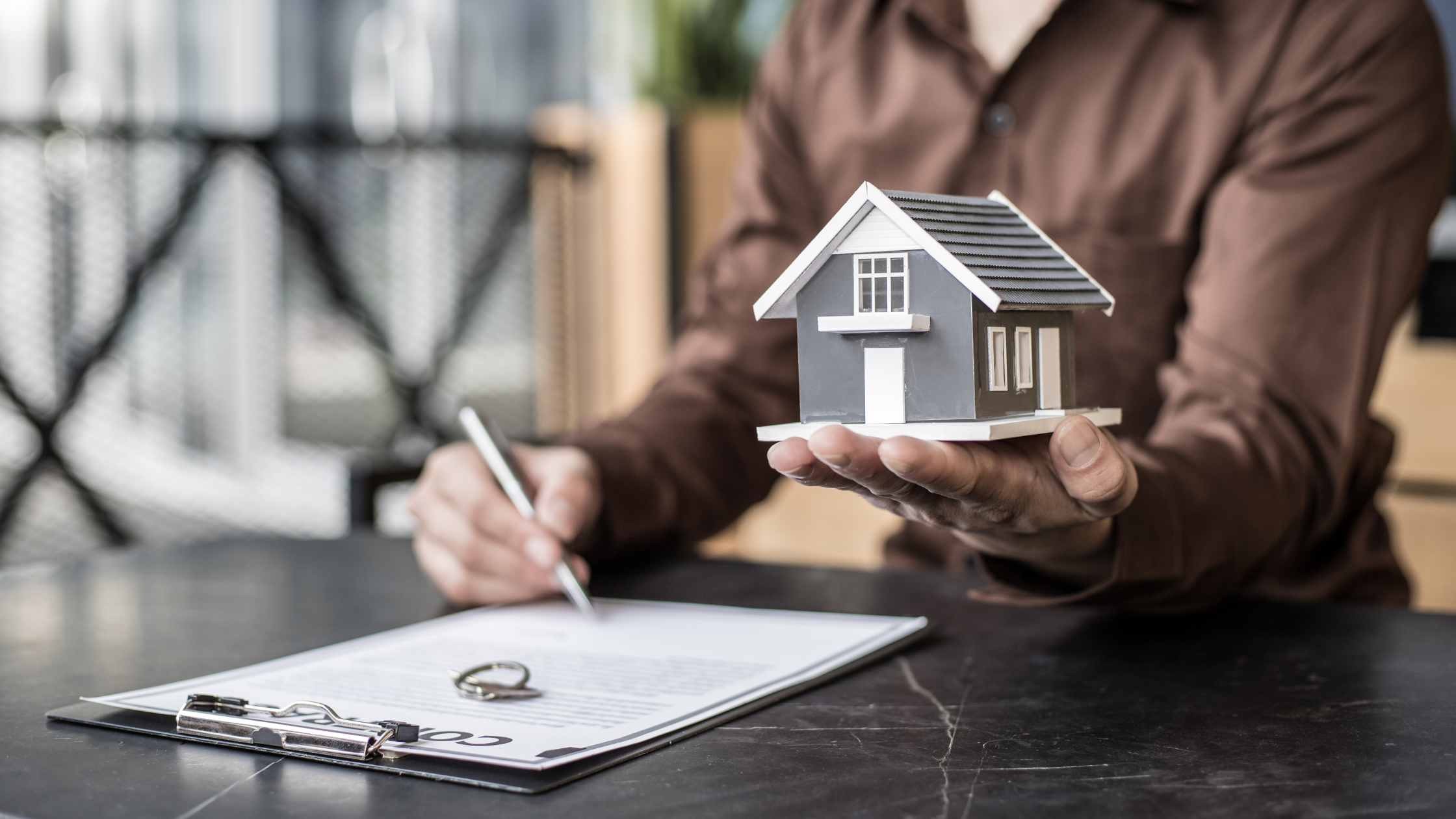 person holding little house in one hand and a pen in the other ready to sign paper work - adam tobin